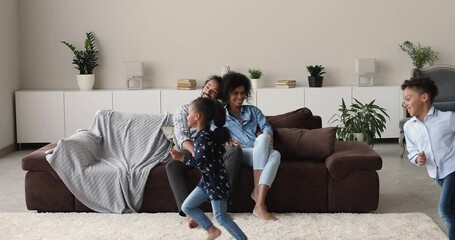 Wall Mural - African 30s couple sit on sofa look at preschool son and daughter running around. Happy spouses and children spend weekend at new house, bank loan for young family, leisure at home with kids concept