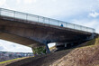 © Dolores  Harvey - An underneath view of a cement overpass bridge with metal rails. The highway structure has grass on the banks and a walking trail is underneath. There's a blue sky with trees in the background.