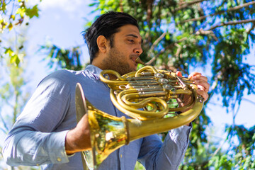 latin man playing a french horn in the park while rehearsing with sheet music. low angle view