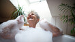 Cheerful senior woman washing in bubble bath tub at home, laughing.