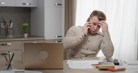 man working at home sitting at his desk talking on the phone unpleasant conversation upset holding h