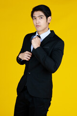 Asian smart man in formal black suit, hand touching tie. Stand and side look to camera. Studio shoot yellow background. Concept modern executive boss.