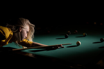 Young attractive woman with dark frizzy hair hitting ball with cue playing billiards. Pool game background