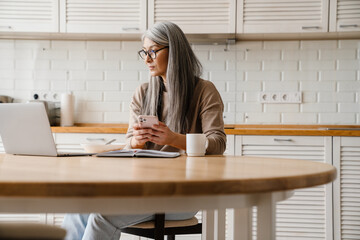 Canvas Print - Mature grey woman working with laptop and cellphone in kitchen