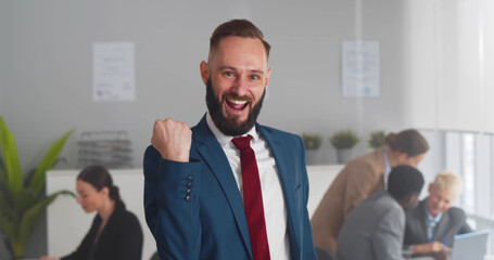 Millennial office worker in suit standing in office raising clenched fists feel overjoyed