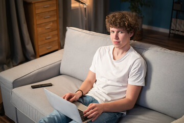 A young boy with curly hair sits on the living room couch with his laptop in his lap and proudly stares into the camera.