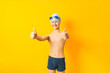 © Olha - School boy dressed in swimming hat, swimming trunks and swimming goggles isolated on yellow background. The child shows a gesture of approx.