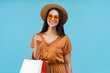 © wpadington - Potrait of beautiful brunette girl with charming smile, posing over pastel blue background in trendy orange yellow sunglasses and boater hat with several shopper bags in hand