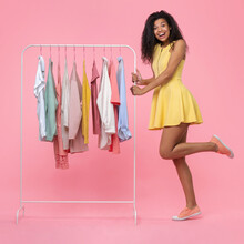 Full Length Portrait Of Cute Happy Smiling Euphoric Dark Skinned Girl Wearing Illuminating Yellow Mini Dress Choosing Clothes Wear Hanging On White Rack In Showroom Isolated On Pastel Pink Background