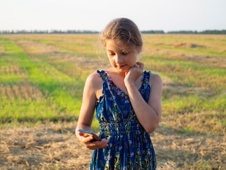 A teenage girl stands in a harvested wheat field and uses her phone to send a message to her friends online. A girl on a green and yellow field in the rays of the setting sun with a phone in her hands