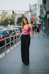 Sticker - Vertical shot of a young blonde female model posing on the sidewalk in a pink blouse and black pants