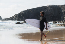 Young Surfer Walking By The Beach Shore Watching The Waves Holding Surfboard