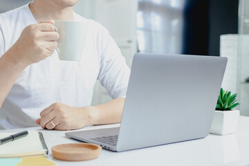 Wall Mural - Man in a white shirt picks up a coffee mug and thinks of work on his laptop while working at home.