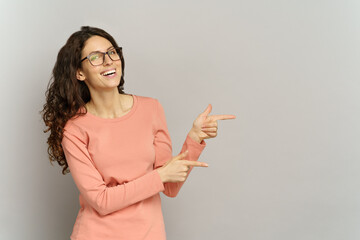 Curly woman in 30s point fingers away to copy space with happy excited smile over gray wall background. Isolated studio portrait of millennial businesswoman in glasses showing presenting displaying