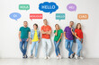 Happy people posing near light wall and illustration of speech bubbles with word Hello written in different languages