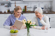 smiling nurse reading book to happy senior woman near fresh apples and tulips in kitchen