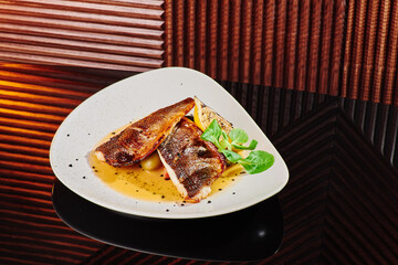 White steak of grilled fish on pillow of vegetables and yellow sauce, with lemon slice and fresh herbs. Fish on white plate on dark beautiful background. Close-up. Space