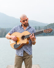 Bearded Guy In Casual Clothes Standing With Guitar On Wooden Pier Near River With Mountains On Background Under Cloudy Gray Sky In Daytime