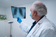 © Enrique Espinosa/ADDICTIVE STOCK - Side view of elderly male doctor in mask and gloves examining x ray image of lungs of patient during coronavirus in hospital