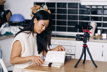 Young Ethnic Female Vlogger With Notebook Sitting At Table With Photo Camera On Tripod In Kitchen