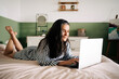 © Jake Jakab/ADDICTIVE STOCK - Side view of positive mature self employed Hispanic woman with long dark hair in casual clothes lying on bed and typing on laptop during online work at home