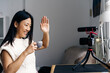 © Jake Jakab/ADDICTIVE STOCK - Smiling ethnic female blogger with cup of hot drink showing hello gesture while recording video on photo camera at home