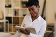 © Jake Jakab/ADDICTIVE STOCK - Cheerful African American female entrepreneur in white shirt standing in room and writing in notepad while working form home and looking at camera