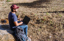 Side view full body of nomad sitting on stone near belongings and using netbook while working remotely