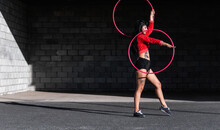 Young Tattooed Woman In Activewear Twirling Hula Hoop While Dancing Against Brick Walls With Shadows And Looking Forward In Sunlight