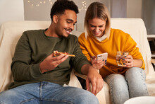 Content Multiracial Couple Sitting In Living Room While Eating Pizza And Watching Funny Video On Smartphone At Weekend