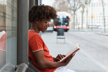 Side View Of Relaxed Young African American Guy With Curly Hair In Casual Clothes And Eyeglasses Reading Book While Waiting For Bus On City Street