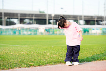 Cute 4 year old Asian girl standing for exercise. Child embraces his arms across the head to opposite side of the head and tilts his neck. Kid enjoy sport. Child wearing pink shirt. Summer or spring.