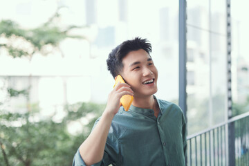 Wall Mural - Asian man talking on smartphone smiling laughing out loud relaxing at balcony