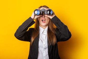 surprised young business woman looking through binoculars on yellow background