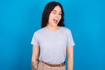 young beautiful tattooed girl wearing blue t-shirt standing against blue background winking looking at the camera with sexy expression, cheerful and happy face.