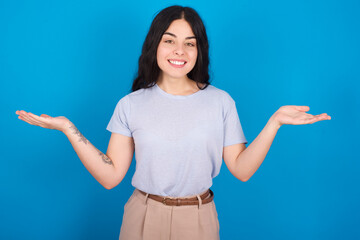 Cheerful cheery optimistic young beautiful tattooed girl wearing blue t-shirt standing against blue background holding two palms copy space