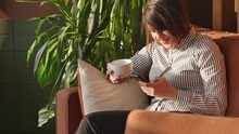 Happy Charming Woman Sitting On Sofa With Cup Of Coffee And Reading Text Message With Her Smartphone During Lunch