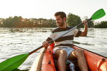 Wall Mural - Excited young man smiling while kayaking in a lake, surrounded by peaceful nature scene on a summer afternoon