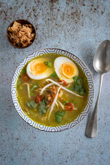 Wall Mural - Top down view of a single bowl of Soto Ayam (translation: Indonesian chicken soup) with a spoon on a textured blue background