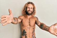 Handsome Man With Beard And Long Hair Standing Shirtless Showing Tattoos Looking At The Camera Smiling With Open Arms For Hug. Cheerful Expression Embracing Happiness.