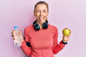 Canvas Print - Beautiful caucasian sports woman holding water bottle and green apple sticking tongue out happy with funny expression.