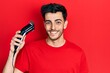 © Krakenimages.com - Young hispanic man charging electric razor machine looking positive and happy standing and smiling with a confident smile showing teeth