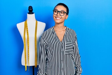 Wall Mural - Beautiful hispanic woman with short hair standing by manikin looking away to side with smile on face, natural expression. laughing confident.