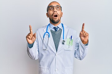 Wall Mural - Hispanic adult man wearing doctor uniform and stethoscope amazed and surprised looking up and pointing with fingers and raised arms.