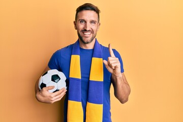 Wall Mural - Handsome man with beard football hooligan cheering game holding ball smiling with an idea or question pointing finger with happy face, number one