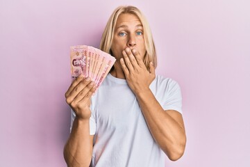 Sticker - Caucasian young man with long hair holding 100 thai baht banknotes covering mouth with hand, shocked and afraid for mistake. surprised expression