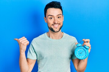 Young hispanic man holding alarm clock pointing thumb up to the side smiling happy with open mouth