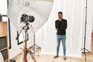 Wall Mural - Young hispanic man with beard posing as model at photography studio looking confident at the camera smiling with crossed arms and hand raised on chin. thinking positive.