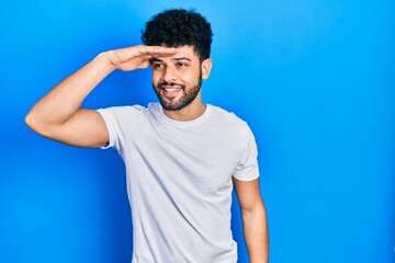 Young arab man with beard wearing casual white t shirt very happy and smiling looking far away with hand over head. searching concept.