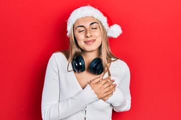 Poster - Beautiful hispanic woman wearing christmas hat and headphones smiling with hands on chest, eyes closed with grateful gesture on face. health concept.